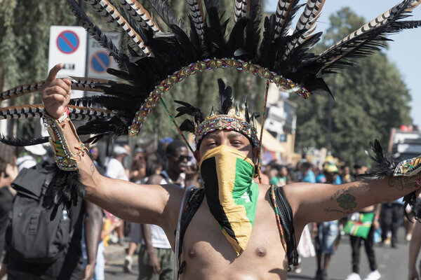 individual in a vibrant costume, including a large feathered headdress and a face covering with Jamaican flag colors, participating in the Notting Hill Carnival in West London, celebrating Caribbean culture and heritage.  the largest street festivals
