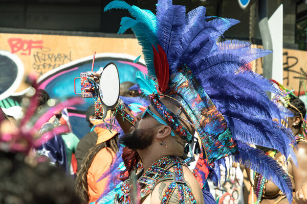 a performer at the Notting Hill Carnival in West London, adorned in a vibrant and intricate costume featuring a large, feathered headdress and ornate jewelry. The Notting Hill Carnival is widely recognized for its celebration of Caribbean culture.