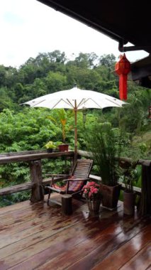 Geleneksel Tayland ev tarzının dikey görüntüleri. Antik mobilyalarla ahşap, dış süslemeler yeşil ağaçlarla süslenmiş ve terasta büyük bir şemsiye.