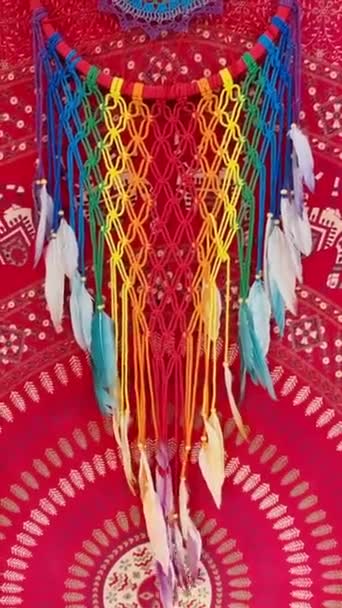 カラフルな夢のキャッチャーの垂直な映像が吊るされ 背景に赤い部族の布が吊るされた風に渦巻きます — ストック動画