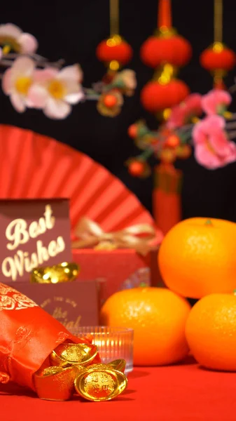 Chinese Lunar New Year background, red theme. gold in silk bag, red gift box with text best wishes, oranges, paper fan, plum blossom branch, candle sway and Chinese new year ornaments for celebrate
