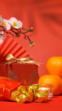 Çin Yeni Yıl kırmızı arka planı. Eski Çin külçeleri ipek çantada altın külçeler, altın yaylı kırmızı hediye kutusu, kağıt yelpaze, portakallar, erik çiçeği dalı, mum ve yaprak gölgesi sallanıyor. dikey