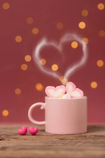 hot drink with heart shape smoke on pink coffee cup with heart shape marshmallow on top, with two small heart placed side by side on wooden table, bokeh light at background. romantic love relationship
