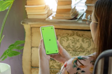 Cep telefonu boş yeşil ekran modeli. Kadın cep telefonunu elinde tutuyor, oturma odasında akıllı telefonu iş bağlantısı ya da eğlence için kullanıyor. Eski dolap, kitap, canavar ağacı ile dekore edilmiş oda.