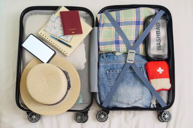 Toparlanmak için yataktaki açık bavul manzarası, yurtdışı seyahatine hazırlanmak pasaport, harita, şapka, seyahat hekimliği ilk yardım çantası, giysi, reklam için fotokopi alanı olan telefon modeli.