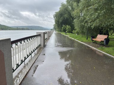 Bulutlu bir günde yağmurdan sonra bir park. Islak bir kırmızı tuğla yolu yemyeşil ağaçlar ve çimenler arasında süzülür. Arka planda banklar ve bir çardak görülebilir.. 