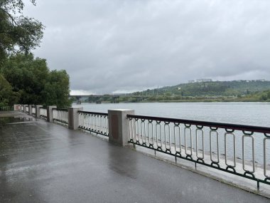 Bulutlu bir günde yağmurdan sonra bir park. Islak bir kırmızı tuğla yolu yemyeşil ağaçlar ve çimenler arasında süzülür. Arka planda banklar ve bir çardak görülebilir.. 