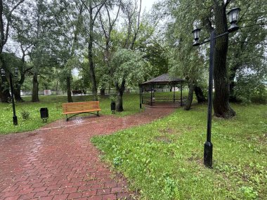 Bulutlu bir günde yağmurdan sonra bir park. Islak bir kırmızı tuğla yolu yemyeşil ağaçlar ve çimenler arasında süzülür. Arka planda banklar ve bir çardak görülebilir.. 