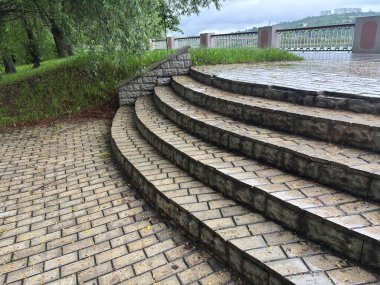 Bulutlu bir günde yağmurdan sonra bir park. Islak bir kırmızı tuğla yolu yemyeşil ağaçlar ve çimenler arasında süzülür. Arka planda banklar ve bir çardak görülebilir.. 