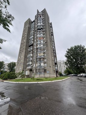 Bulutlu bir günde alçak bir açıdan çekilmiş uzun bir Sovyet döneminden kalma apartman. Bina, bazıları cam veya metal ızgaralarla çevrili ve ağaçlar ve park halindeki arabalarla çevrili çok sayıda balkona sahip..