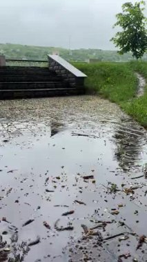 Bulutlu bir günde yağmurdan sonra bir park. Islak bir kırmızı tuğla yolu yemyeşil ağaçlar ve çimenler arasında süzülür. Arka planda banklar ve bir çardak görülebilir.. 