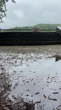 Bulutlu bir günde yağmurdan sonra bir park. Islak bir kırmızı tuğla yolu yemyeşil ağaçlar ve çimenler arasında süzülür. Arka planda banklar ve bir çardak görülebilir.. 