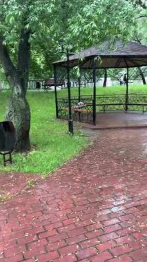 Bulutlu bir günde yağmurdan sonra bir park. Islak bir kırmızı tuğla yolu yemyeşil ağaçlar ve çimenler arasında süzülür. Arka planda banklar ve bir çardak görülebilir.. 