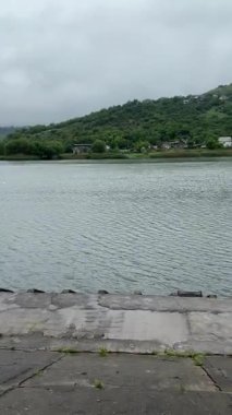 Bulutlu bir günde yağmurdan sonra bir park. Islak bir kırmızı tuğla yolu yemyeşil ağaçlar ve çimenler arasında süzülür. Arka planda banklar ve bir çardak görülebilir.. 