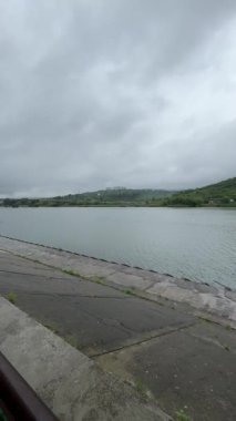 Bulutlu bir günde yağmurdan sonra bir park. Islak bir kırmızı tuğla yolu yemyeşil ağaçlar ve çimenler arasında süzülür. Arka planda banklar ve bir çardak görülebilir.. 