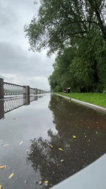 Bulutlu bir günde yağmurdan sonra bir park. Islak bir kırmızı tuğla yolu yemyeşil ağaçlar ve çimenler arasında süzülür. Arka planda banklar ve bir çardak görülebilir.. 