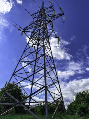 Uzun çelik bir elektrik iletim kulesi, kabarık beyaz bulutlarla kaplı canlı mavi bir gökyüzüne doğru hızla yükseliyor. Kafes kulesinin geometrik yapısı çarpıcı bir karşıtlık oluşturur