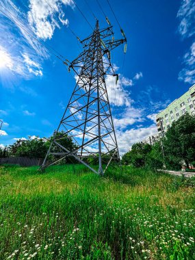 Uzun çelik bir elektrik iletim kulesi, kabarık beyaz bulutlarla kaplı canlı mavi bir gökyüzüne doğru hızla yükseliyor. Kafes kulesinin geometrik yapısı çarpıcı bir karşıtlık oluşturur