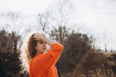 Sonbahar parkının arka planında güzel gülüşler kıvırcık esmer. Sonbahar doğasının arka planına karşı turuncu kazaklı genç kıvırcık kız.