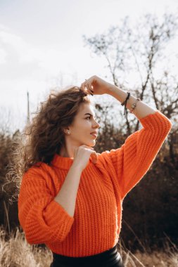 Sonbahar parkının arka planında güzel gülüşler kıvırcık esmer. Sonbahar doğasının arka planına karşı turuncu kazaklı genç kıvırcık kız.