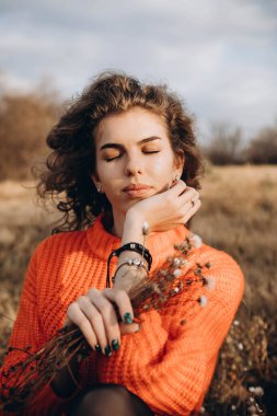 Sonbahar parkının arka planında güzel gülüşler kıvırcık esmer. Sonbahar doğasının arka planına karşı turuncu kazaklı genç kıvırcık kız.