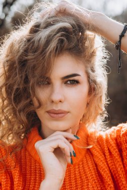 Sonbahar parkının arka planında güzel gülüşler kıvırcık esmer. Sonbahar doğasının arka planına karşı turuncu kazaklı genç kıvırcık kız.