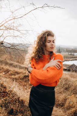 Sonbahar parkının arka planında güzel gülüşler kıvırcık esmer. Sonbahar doğasının arka planına karşı turuncu kazaklı genç kıvırcık kız.