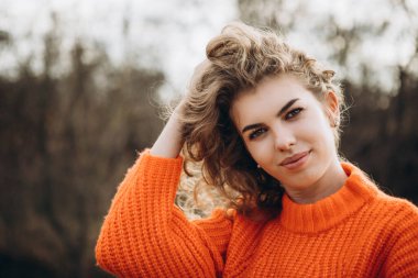 Sonbahar parkının arka planında güzel gülüşler kıvırcık esmer. Sonbahar doğasının arka planına karşı turuncu kazaklı genç kıvırcık kız.