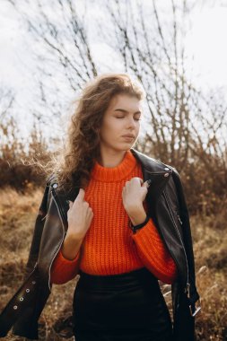 Sonbahar parkının arka planında güzel gülüşler kıvırcık esmer. Sonbahar doğasının arka planına karşı turuncu kazaklı genç kıvırcık kız.