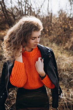 Sonbahar parkının arka planında güzel gülüşler kıvırcık esmer. Sonbahar doğasının arka planına karşı turuncu kazaklı genç kıvırcık kız.