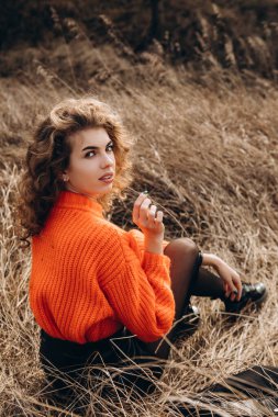 Sonbahar parkının arka planında güzel gülüşler kıvırcık esmer. Sonbahar doğasının arka planına karşı turuncu kazaklı genç kıvırcık kız.