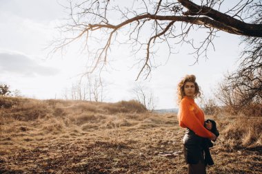 Sonbahar parkının arka planında güzel gülüşler kıvırcık esmer. Sonbahar doğasının arka planına karşı turuncu kazaklı genç kıvırcık kız.