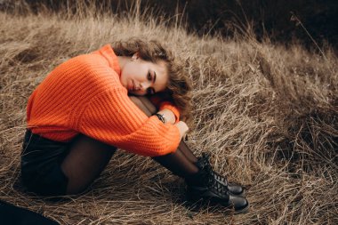 Sonbahar parkının arka planında güzel gülüşler kıvırcık esmer. Sonbahar doğasının arka planına karşı turuncu kazaklı genç kıvırcık kız.