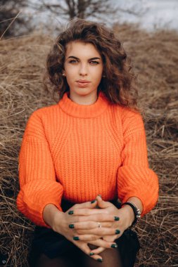 Sonbahar parkının arka planında güzel gülüşler kıvırcık esmer. Sonbahar doğasının arka planına karşı turuncu kazaklı genç kıvırcık kız.