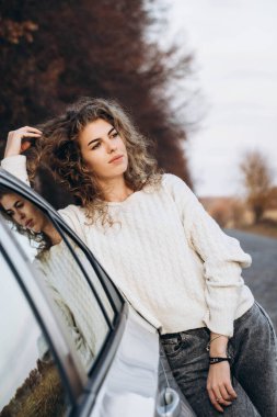 Sonbahar doğasının ve kendi arabasının arka planına karşı beyaz kazaklı genç kıvırcık kız. Yüksek kalite fotoğraf