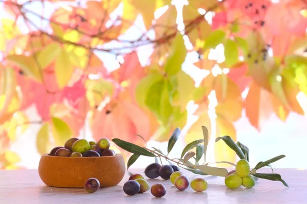 Olives on wooden table in the garden, olive oil in glass jug and autumn leaves background