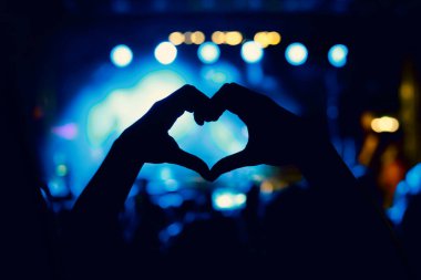 A person is making a heart sign during a concert of a favorite music band. Black silhouette
