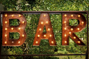 Parlak harfler. Paslı metalden yükselen lambalardan oluşan bir çelenk ile gelen ses metrik harfler.