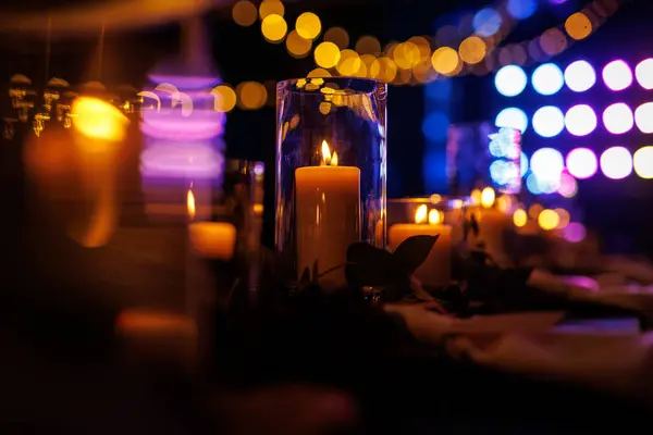 A romantic table setting with lit candles and greenery at a nighttime event. The candles create a warm and intimate atmosphere, while the greenery adds a touch of elegance to the setting.
