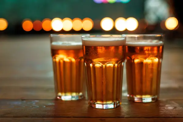 Three pints of frothy beer on a wood table with a blurred party backdrop.