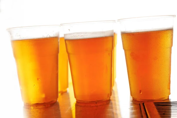 Plastic glasses of frothy beer on a metal table with a white backdrop.