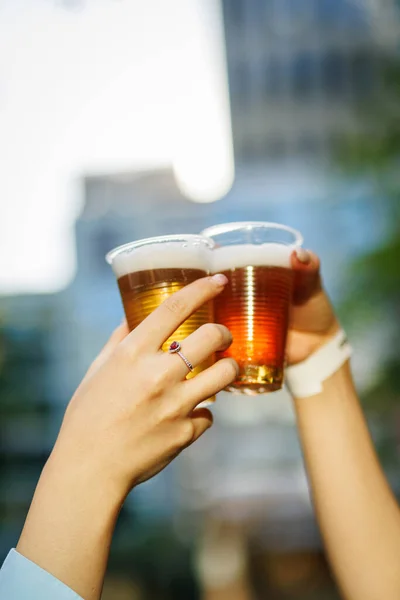 A pair of hands cheers with beer glasses in a blurred sunset background.