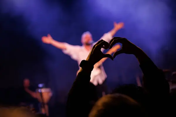 The concert music fan make a heart shape with their hands. Happy singer with raised hands in the background