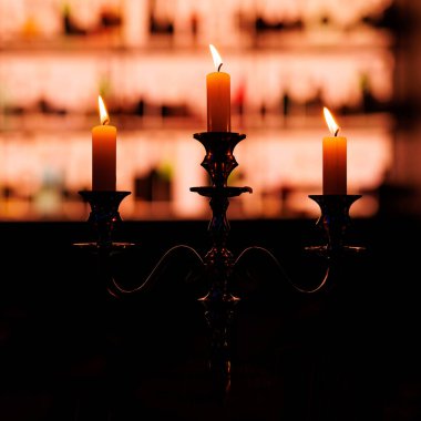 Üç yanan mumla birlikte eski bir şamdan, aydınlanmış raflardan oluşan bir zemin üzerine sıcak bir parıltı ve siluet konur..