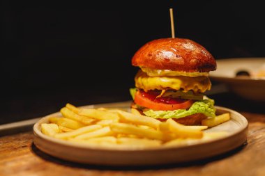 Lezzetli bir hamburger, sulu bir köfte, erimiş peynir, taze marul ve domates dilimleri ile istiflenmiş, ahşap bir tabakta çıtır altın patates kızartması ile birlikte servis edilen.