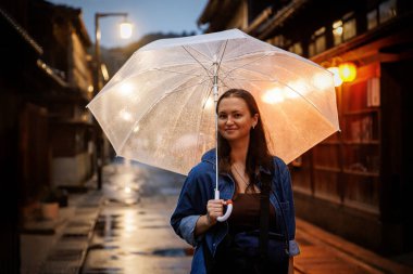 Gün batımında yağmurlu bir sokakta bir şemsiyeyi zarif bir şekilde tutan bir kadının huzur yarattığı huzurlu ve güzel bir sahne.