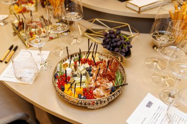Decorative platter with assorted cheese, meats, herbs and berries. Surrounded by empty wine glasses.