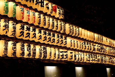 Açık havada asılı duran aydınlatılmış Japon kağıt fenerleri. Kalın siyah kanji yazısıyla süslenmiş..
