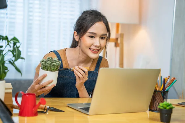 Japanese Asian female blogger holding a pot of plants watering and shoveling the soil, reviewing planting online with a laptop.