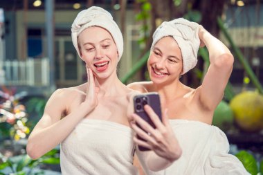 Two Caucasian female friends in towel and head wrap resting relaxing massage in spa, happy friends together taking smartphone selfie in hotel spa salon.wellness, beauty ,spa and relaxation concept